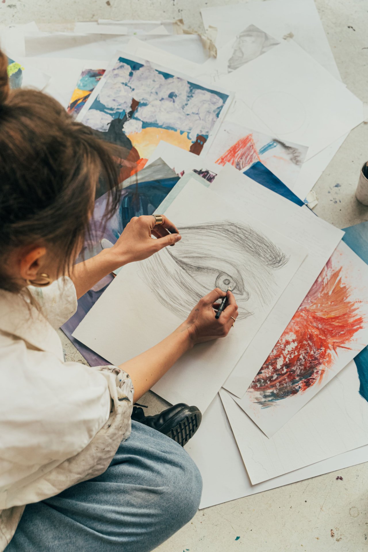 Artista a desenhar um retrato a lápis rodeada de pinturas coloridas no chão do atelier
